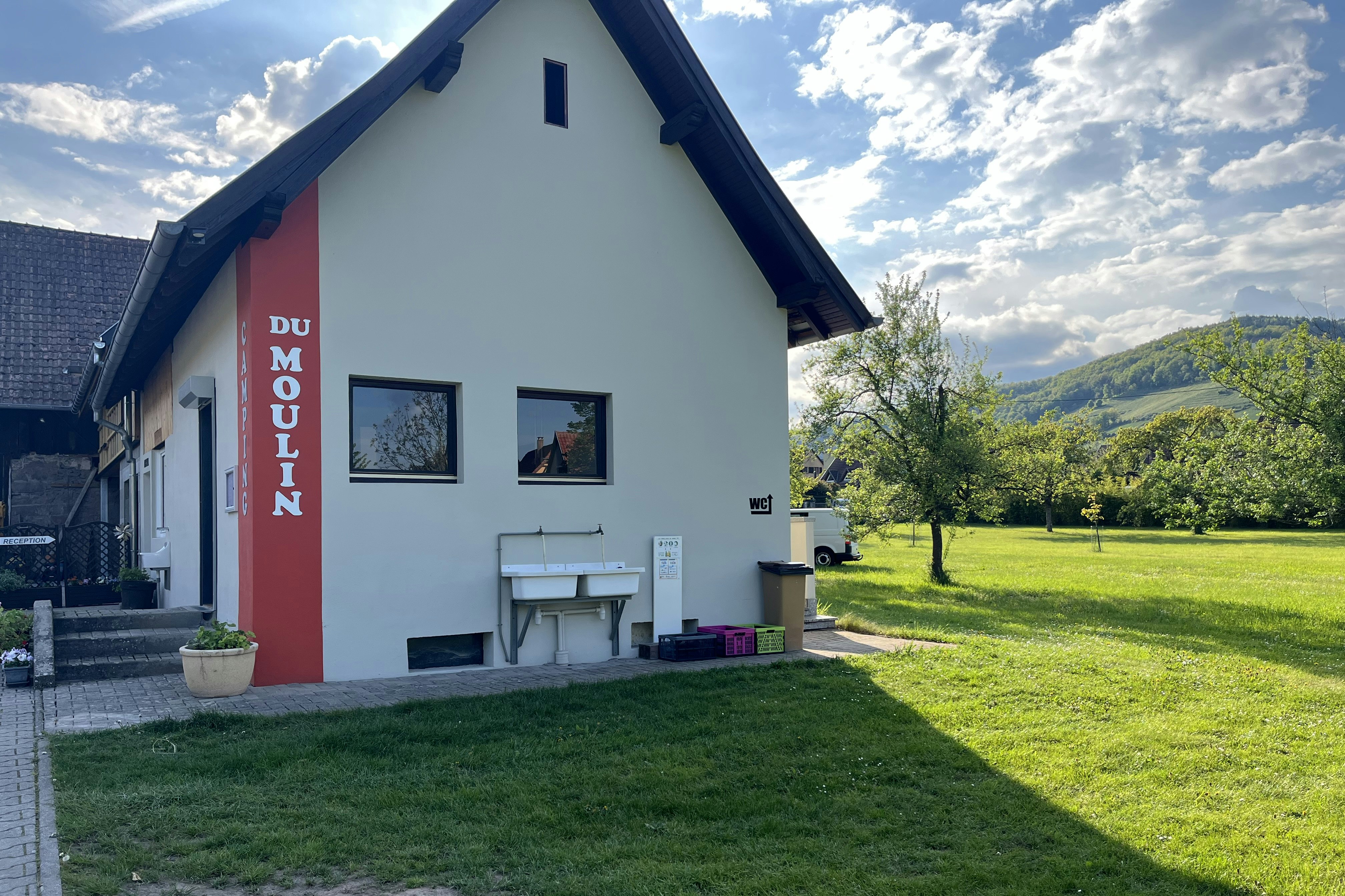 Weiße Ferienhaus-Fassade mit rotem Eingang, „DU MOLIN“ am Schild, Tisch und Stühlen vor dem Haus, Rasen und Bäume im Garten.