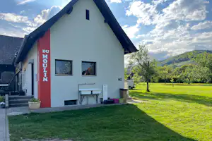 Weiße Ferienhaus-Fassade mit rotem Eingang, „DU MOLIN“ am Schild, Tisch und Stühlen vor dem Haus, Rasen und Bäume im Garten.