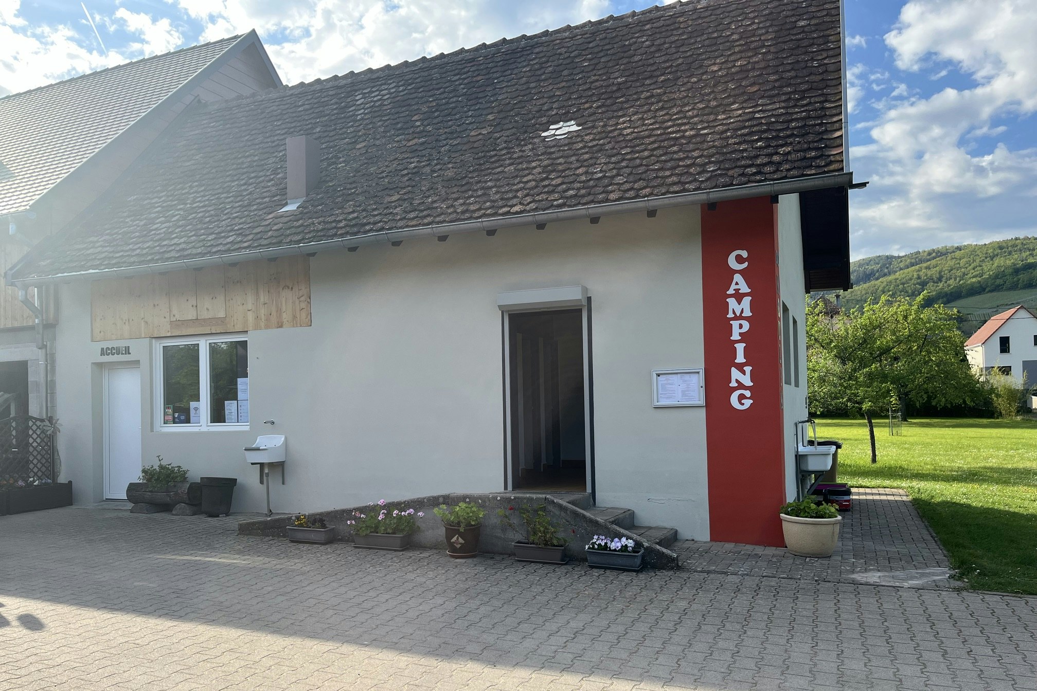Campinggebäude mit Eingangstür und rotem Säulenbanner „CAMPING“, davor gepflasterter Weg und Rasenfläche mit Hecke.