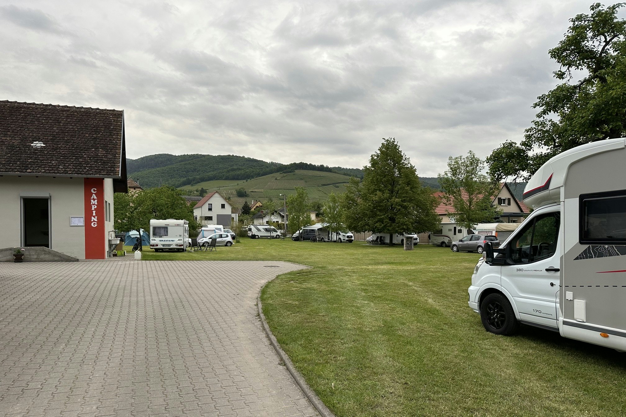 Wohnmobil und Stellplätze auf einem Campingplatz, gepflasterter Weg, Rasenfläche und Bäume vor Häusern im Hintergrund.