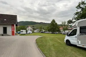 Wohnmobil und Stellplätze auf einem Campingplatz, gepflasterter Weg, Rasenfläche und Bäume vor Häusern im Hintergrund.