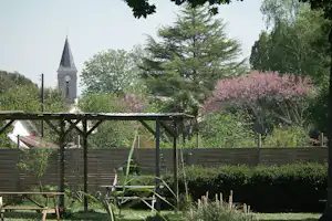 Blick vom Campinggarten auf einen Kirchturm mit Uhr, umgeben von Bäumen und blühenden Fliedersträuchern.