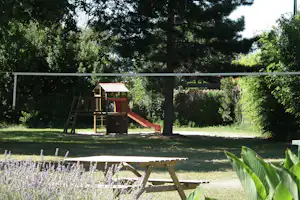 Spielplatz mit hölzernem Kletterturm und roter Rutsche im Grünen, umgeben von Bäumen und Rasen.