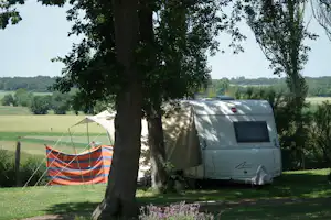 Wohnwagen und Zelt am Campingplatz unter großen Bäumen, mit Wiese und Feldlandschaft im Hintergrund.