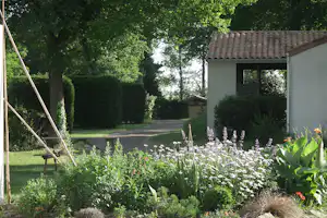 Gartenweg zwischen Bäumen und Hecken, im Vordergrund Blumenbeete, rechts ein Gebäude mit offenem Durchgang.