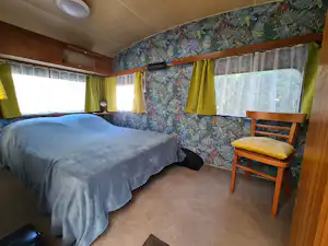 Schlafzimmer in einem Wohnwagen mit Doppelbett, Holzverkleidung, gemusterten Tapeten und gelben Vorhängen vor den Fenstern.