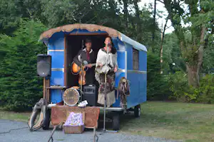 Blauer Camper mit Musikern und Gitarren auf einem Campingplatzweg, davor Gepäck und Deko, im Hintergrund Bäume.