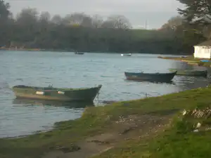 Ruderboote am Ufer eines Sees, grasiger Weg am Campingplatz und ein kleines Bootshaus am Rand.