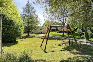 Spielplatz mit zwei Schaukeln auf Rasen, daneben ein Wohnmobil und ein Weg zwischen Bäumen.