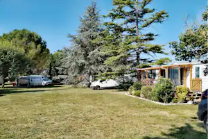 Wohnwagen und Auto auf einer grünen Campingwiese, daneben ein Mobilheim mit Holzveranda und Geländer, umgeben von Bäumen.