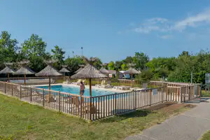 Schwimmbecken auf dem Campingplatz mit Umzäunung, Liegen und Sonnenschirmen im Hintergrund, Rasen und Weg am Beckenrand.