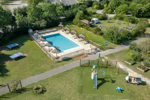 Campingplatz mit beheiztem Pool im umzäunten Bereich, Liegen und Rasenflächen, umgeben von Bäumen und Wegen.