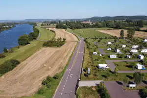 Blick auf einen Campingplatz mit Stellplätzen, Wohnmobilen und einem breiten Weg entlang eines Flusses und grüner Felder.