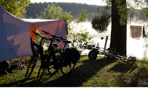 Campingzelt am Waldrand, davor Fahrräder und ein Anhänger auf dem Stellplatz, im Hintergrund Bäume und Hügel.