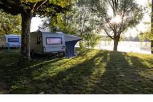 Wohnwagen mit Vorzelt am Campingplatz im Grünen, Bäume und Rasenfläche im Vordergrund, Sonnenlicht durch die Blätter.