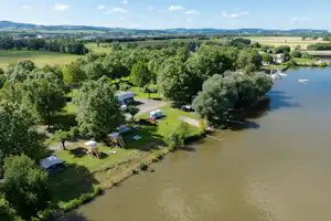 Luftaufnahme eines Campingplatzes am Fluss mit Stellplätzen, Zelten, Tischen und Bäumen entlang des Ufers.