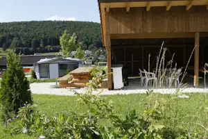 Holzgebäude mit überdachter Terrasse, daneben Wohnwagen und Kiesfläche, im Hintergrund Bäume und Hügel.