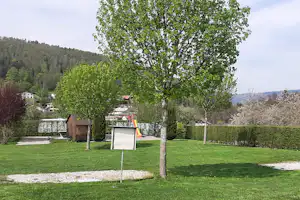 Grüner Campingplatz mit Rasen, Baum, Infotafel und Hecke; im Hintergrund Gebäude und bewaldete Hügel.