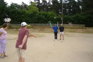 Männer spielen Boule auf einer Sandfläche im Campingpark, im Hintergrund Bäume und ein Holzgeländer.