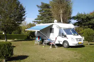 Weißes Wohnmobil mit Markise auf dem Stellplatz, Campingstühle und Tisch vor dem Fahrzeug, umgeben von Bäumen und Hecken.