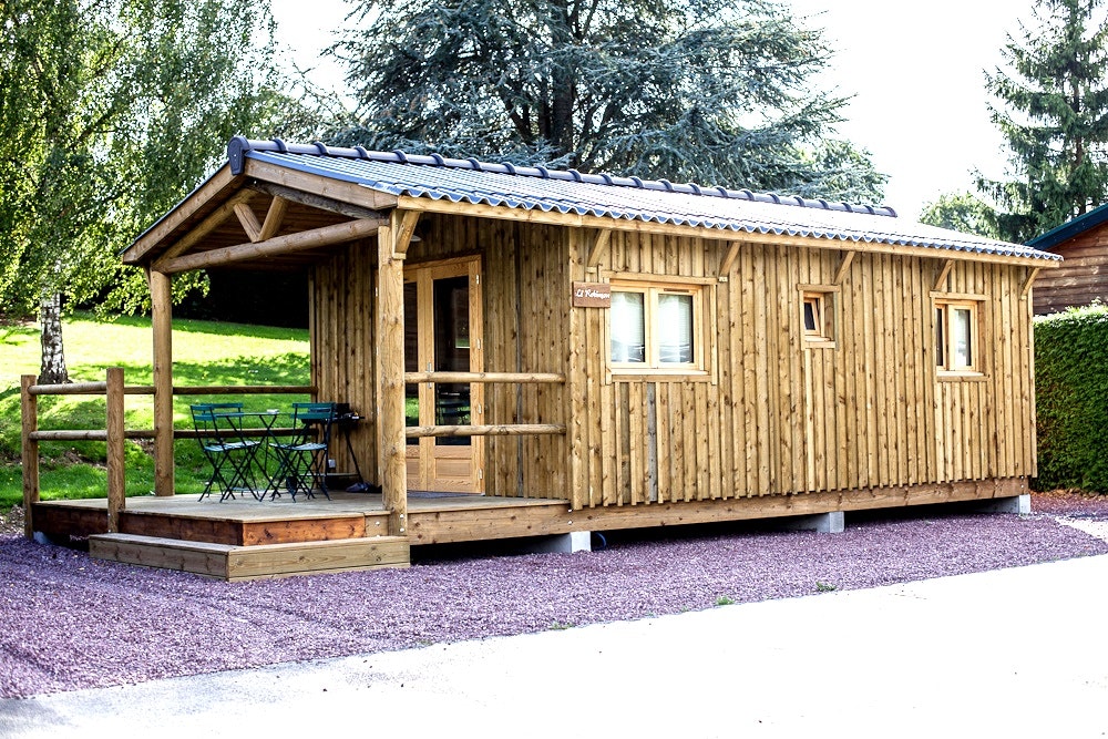 Holzhütte mit Veranda, Sitzbank und Tisch auf Kiesstellplatz, umgeben von Bäumen und grünem Rasen.