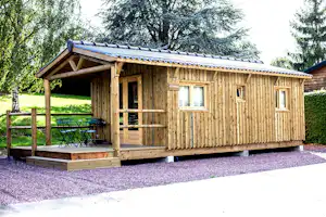 Holzhütte mit Veranda, Sitzbank und Tisch auf Kiesstellplatz, umgeben von Bäumen und grünem Rasen.