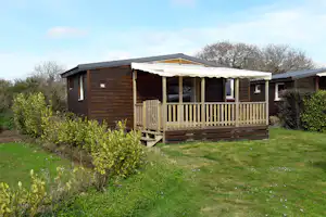 Holzbungalow mit Veranda und Markise, umgeben von Rasen, Hecken und Büschen vor weiteren Gebäuden.