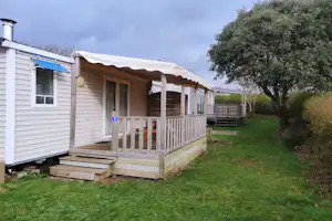 Holz-Mobilheim mit Veranda und Markise, Stufen zum Eingang, Rasenfläche und Bäume am Campingplatz.