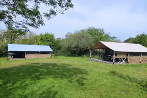 Campingwiese mit zwei überdachten Bereichen, Holzhütten und Tischen, umgeben von Bäumen und grünem Rasen.