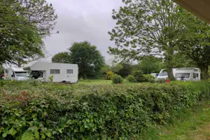Wohnwagen und Wohnmobile auf einem Campingplatz, hinter einer Hecke mit Bäumen und Rasenfläche.