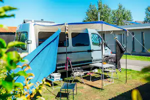 Wohnmobil mit blauem Vorzelt, Tisch und Campingstühlen am Stellplatz, im Hintergrund Gebäude und Bäume.