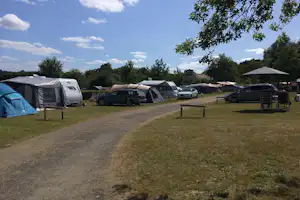 Campingplatz mit Stellplätzen, Zelten und Wohnwagen, Autos am Weg, Rasenfläche und Bäume im Hintergrund.