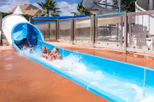 Blaue Wasserrutsche mit Kindern im Rutschbereich, Holzgeländer und Sonnenschirme am Campingplatz, im Hintergrund Gebäude und Palmen.
