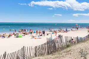 Strand mit vielen Badegästen, Liegestühlen und Stranddecken, im Hintergrund das Meer und ein Zaun aus Holzpfählen.