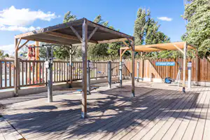 Fitnessgeräte auf einer Holzterrasse unter einem großen Holzvordach, mit Zaun und Bäumen im Hintergrund.