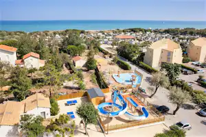 Blick auf einen Campingplatz mit Pool, Sonnenterrasse, Wohngebäuden und Bäumen, nahe dem Meer am Horizont.