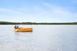 Gelbes Schlauchboot mit zwei Personen auf einem See, im Hintergrund bewaldete Uferlinie und blauer Himmel.