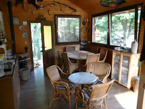 Essbereich in einer Holzhuette mit Esstisch, Korbstuehlen, grossen Fenstern und Tueren zur Terrasse im Gruen.