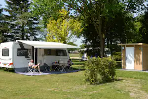 Wohnwagen mit Vorzelt und Sitzgruppe auf dem Campingplatz, daneben ein weiteres Fahrzeug und ein Garten mit Bäumen.