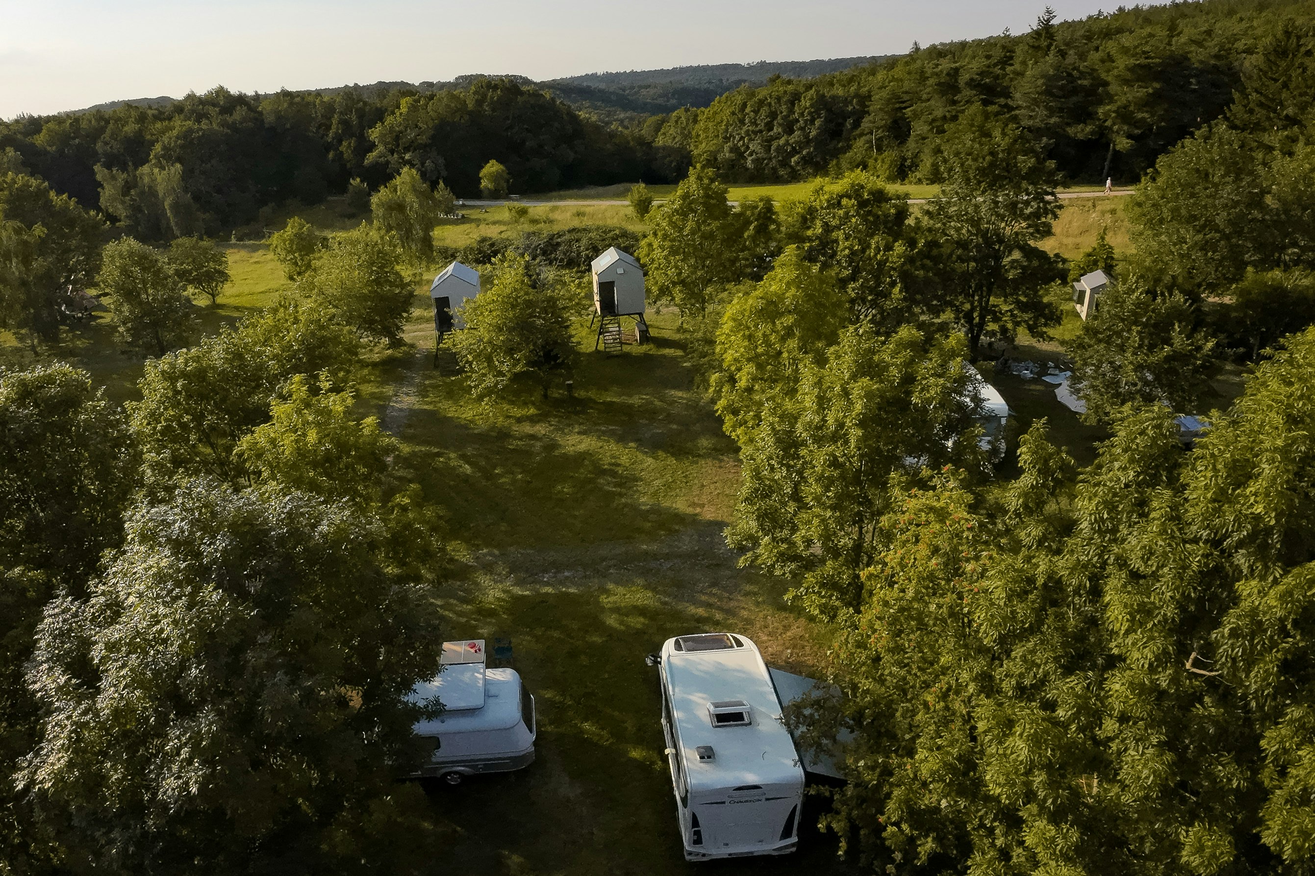 Campingplatz im Grünen: zwei Wohnwagen auf Stellplätzen, umgeben von Bäumen, mit kleinen Hütten im Hintergrund.