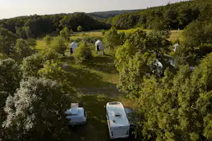 Campingplatz im Grünen: zwei Wohnwagen auf Stellplätzen, umgeben von Bäumen, mit kleinen Hütten im Hintergrund.