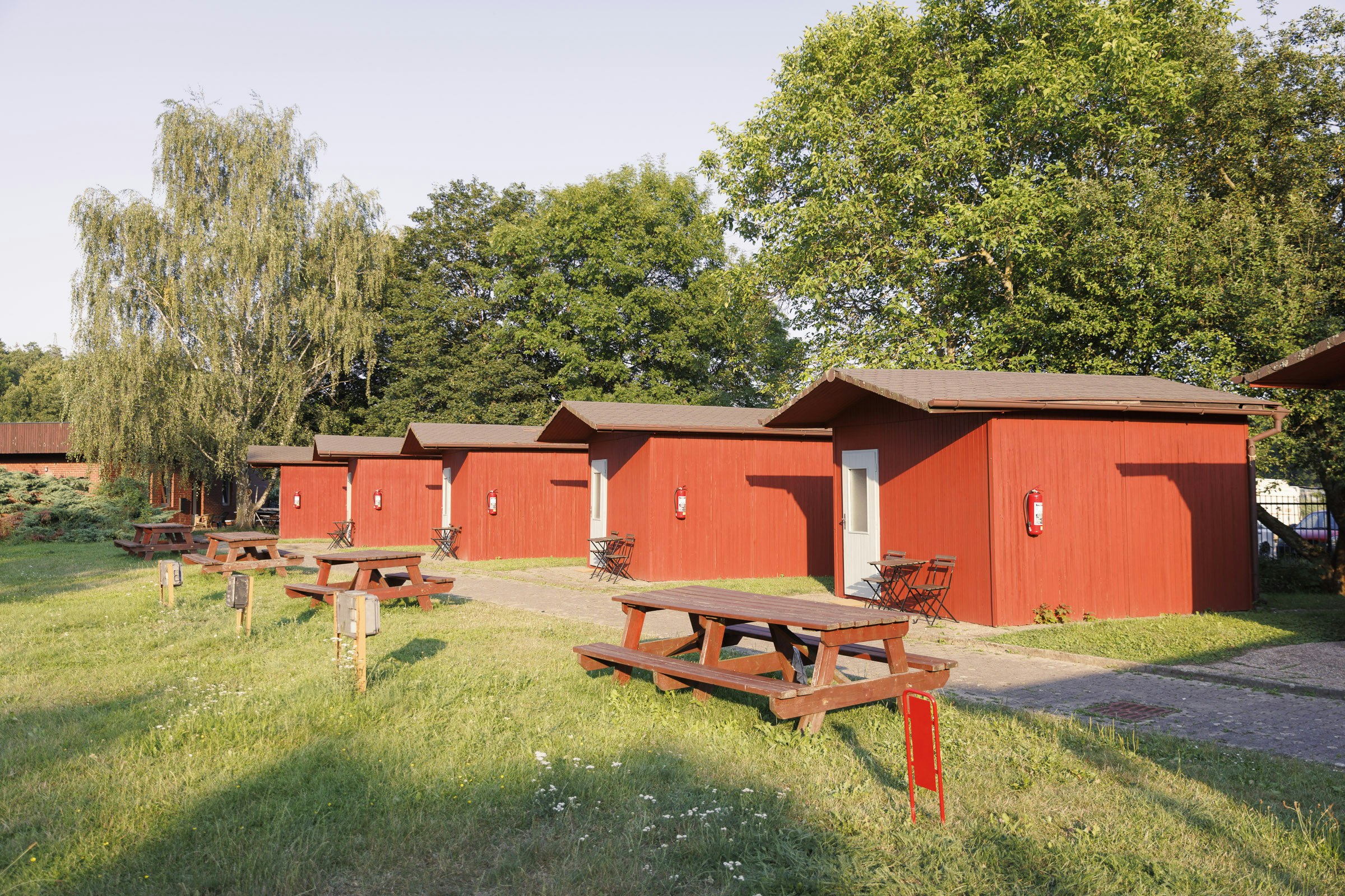 Rote Campinghütten mit Holz-Tischen und Bänken auf einer grünen Wiese, daneben ein gepflasterter Weg und Bäume im Hintergrund.