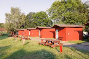 Rote Campinghütten mit Holz-Tischen und Bänken auf einer grünen Wiese, daneben ein gepflasterter Weg und Bäume im Hintergrund.