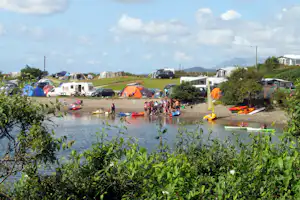 Campingplatz am See: Zelte, Wohnwagen und Autos am Ufer, Kajaks im Wasser und viele Menschen am Strandbereich.