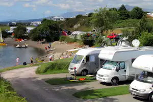 Wohnmobile und Zelte auf einem Campingplatz am Fluss, mit Rasen, Wegen und Bäumen am Hang.