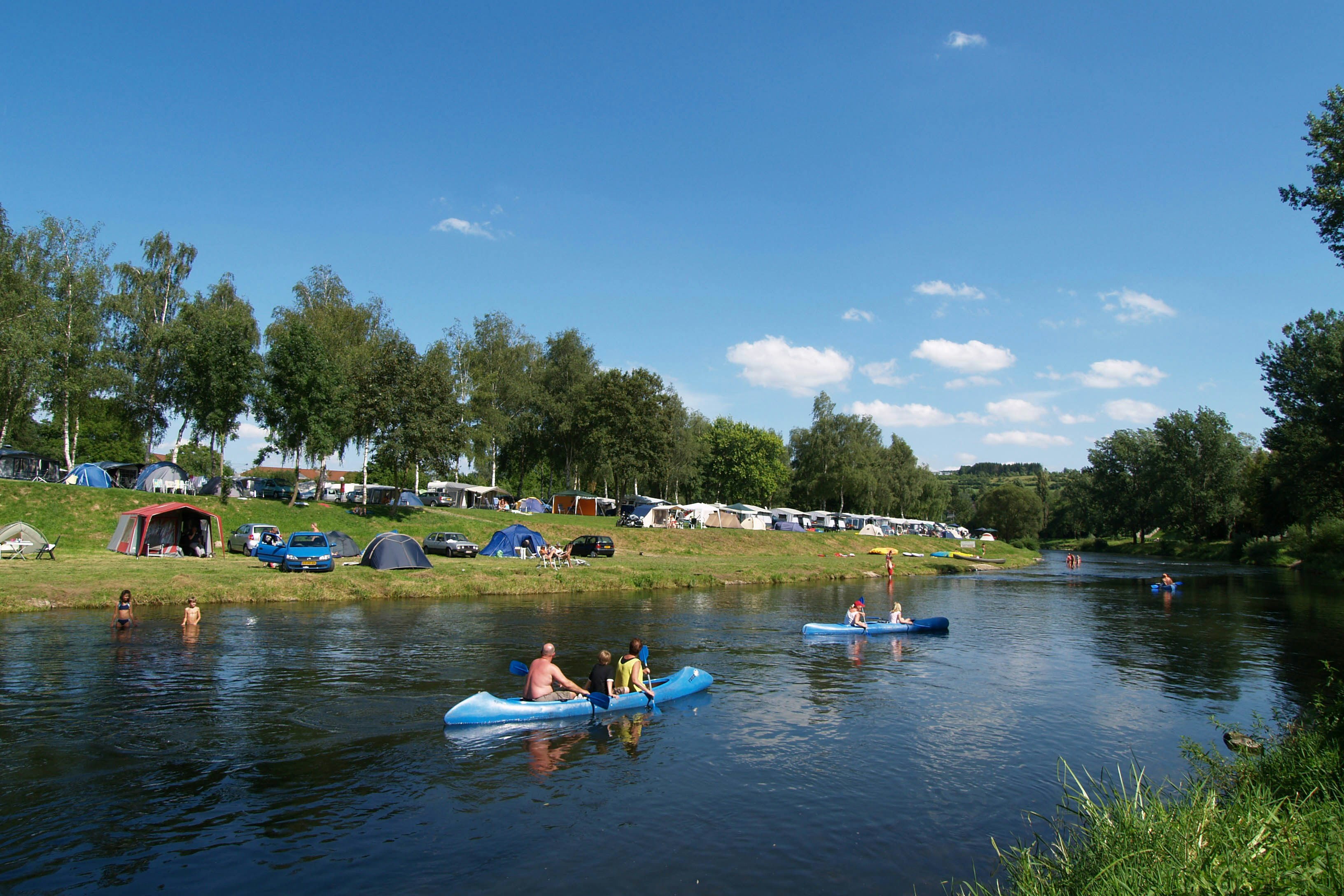 Campingplatz am Fluss: Zelte und Autos auf der Wiese, Menschen im Wasser auf blauen Schlauchbooten.