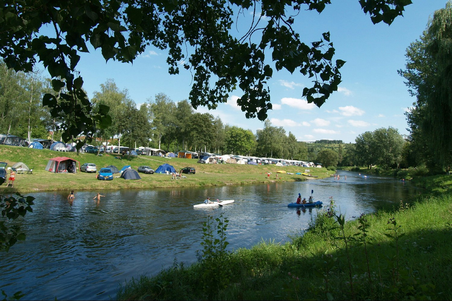 Campingplatz am Fluss mit Zelten und Wohnwagen am Ufer, Ruderboot und Kanus auf dem Wasser, Bäume im Hintergrund.