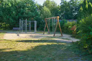 Spielplatz auf dem Campingplatz mit Sandfläche, Klettergerüst und eingezäuntem Bereich, umgeben von Bäumen und Grün.