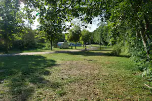 Campingplatz mit Rasenfläche und Bäumen, im Hintergrund ein Wohnwagen und ein Wohnmobil auf dem Stellplatz.