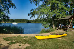 Gelbes Kajak am Ufer am Campingplatz, Grasfläche und Bäume am See mit Booten in der Ferne.